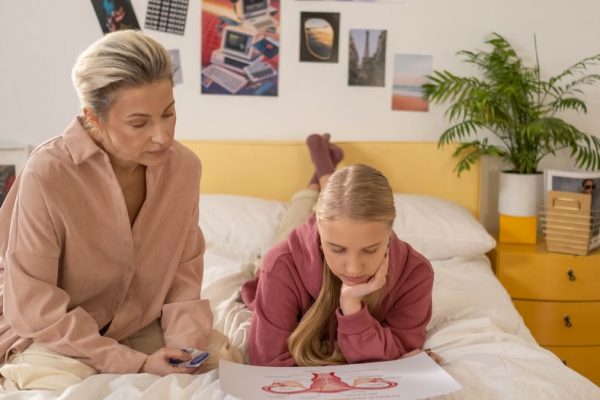 A mother and daughter talk over illustrated materials in a cozy bedroom setting.